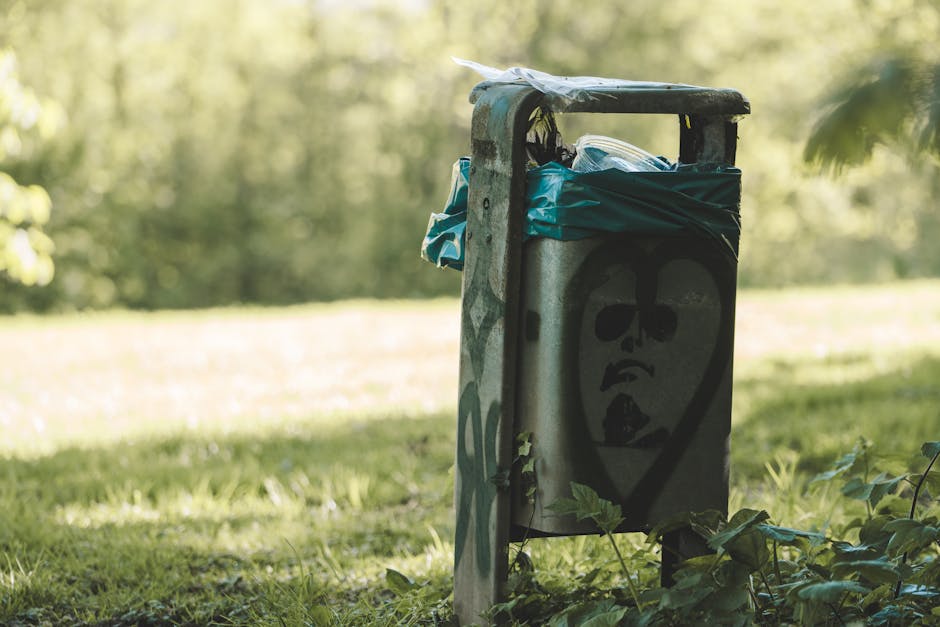 A metal rubbish bin situated outdoors on a grassy area with a blurred natural background of trees and sunlight. The bin features a graffiti-style face painted in black on its front panel, which has a metallic, slightly weathered finish. The top of the bin is open, revealing a blue plastic bag spilling slightly over the edges, containing waste materials. The bin is positioned near some low green foliage, and the environment appears to be a park or garden setting. The scene is illuminated with natural daylight, highlighting the rough texture of the metal surface and the contrast between the graffiti artwork and the surrounding greenery, reflecting a typical scene of informal waste disposal or illegal dumping outside of private or public waste collection points.