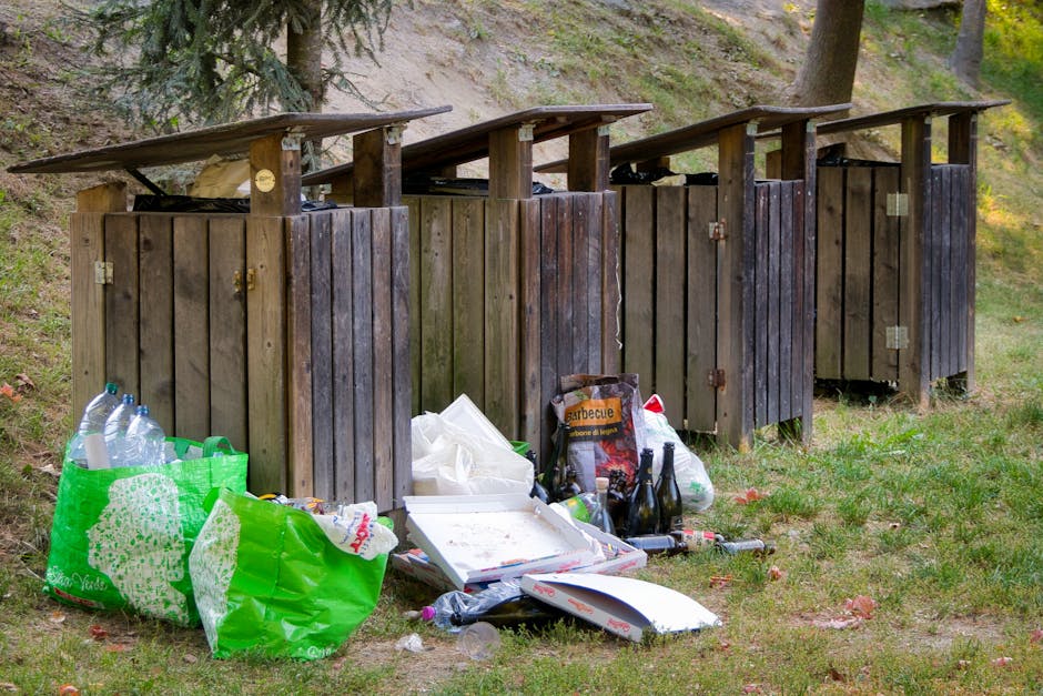 A bright green, dome-shaped public rubbish bin with a semi-transparent black plastic top, filled with mixed waste including crumpled paper and plastic wrappers. The bin features a blue plastic bag lining the interior, secured around the opening. It is mounted on a metal pole set on a paved area with grass, dirt, and a natural landscape in the background, illuminated by natural daylight. The scene suggests this waste receptacle aims to promote proper disposal in an outdoor environment, aligning with private waste handling or alternative rubbish collection services such as those provided by wastecollectionfinsburypark.co.uk for efficient rubbish removal and on-site clearance.