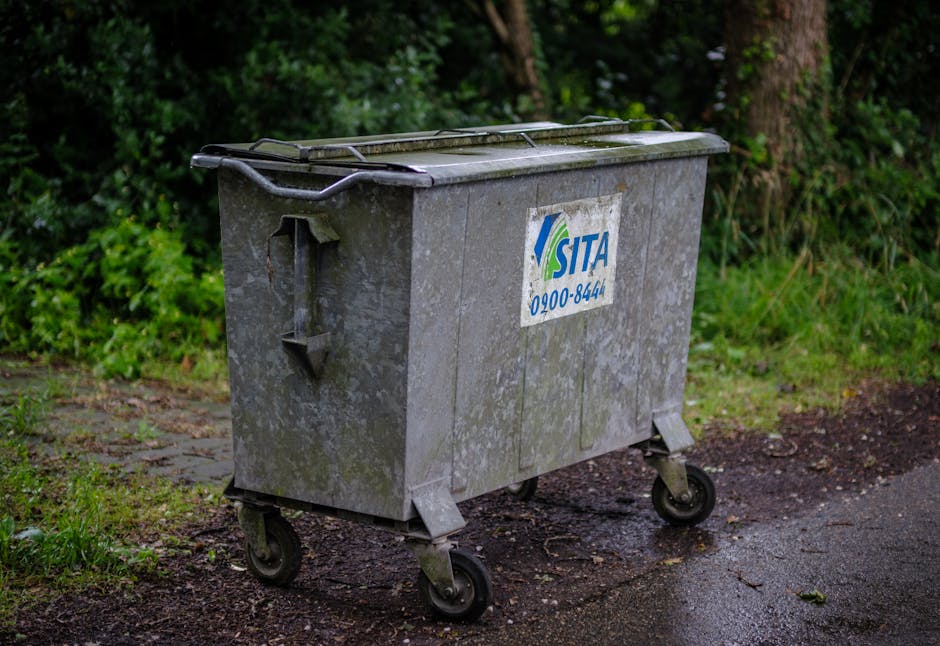 A large, rectangular metal waste container with a weathered, grey finish, positioned on a paved outdoor surface. The bin has a closed lid with a slightly raised, textured surface, showing signs of dirt and moss accumulation. It is mounted on four small black wheels and features a blue and white label with the company logo and contact number, partially worn but still legible. The container is situated beside a rough, paved path with some patches of mud and dirt, bordered by green foliage and trees, indicating an outdoor setting likely at the edge of a residential or park area. The scene is illuminated by natural daylight, highlighting the utilitarian appearance of the bin and its surroundings, consistent with a private waste collection or rubbish removal service, such as those provided by independent waste management companies.