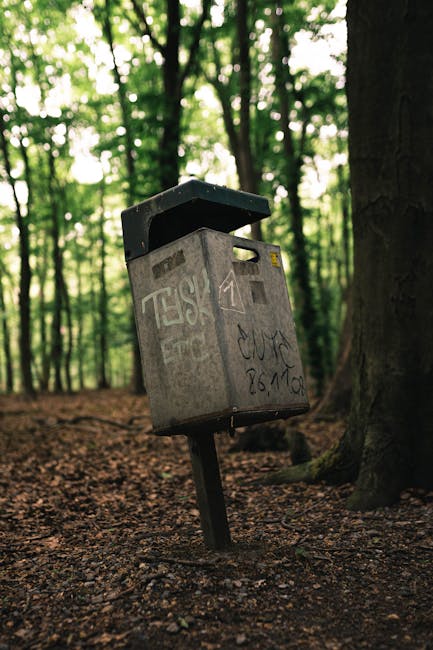A weathered grey metal public mailbox with a partially open black lid, situated in a wooded area on a dirt ground covered with fallen leaves. The mailbox has visible graffiti marks and scratches on its surface, leaning slightly to one side and mounted on a single black metal post embedded in the ground. Surrounding the mailbox are tall trees with dark trunks and green foliage, with dappled sunlight filtering through the leaves, creating a natural, outdoor environment. The scene suggests a rural or park setting, contrasting with typical urban rubbish collection points, and subtly relates to alternative waste handling in outdoor spaces or private collection contexts. The overall scene remains neutral and factual, without promotional language, aligning with waste management and rubbish removal themes.
