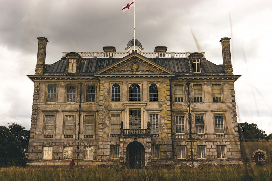 A large, historic stone manor house with a symmetrical façade featuring three stories, numerous rectangular sash windows with white frames, and a central balcony above the main entrance. The building's exterior shows weathered stonework with visible discoloration and moss growth, indicating age and exposure to the elements. The roof is made of dark slate tiles, with multiple chimneys and a small central dome with a Union Jack flag flying on top. The scene is outdoors, set against a cloudy sky with overcast lighting, and the manor is surrounded by overgrown grass and trees in the background, suggesting a private estate or historical site. This detailed description aligns with the context of property clearance or on-site waste disposal activities often associated with private or estate rubbish removal services, with no visible rubbish or waste objects in the image itself.