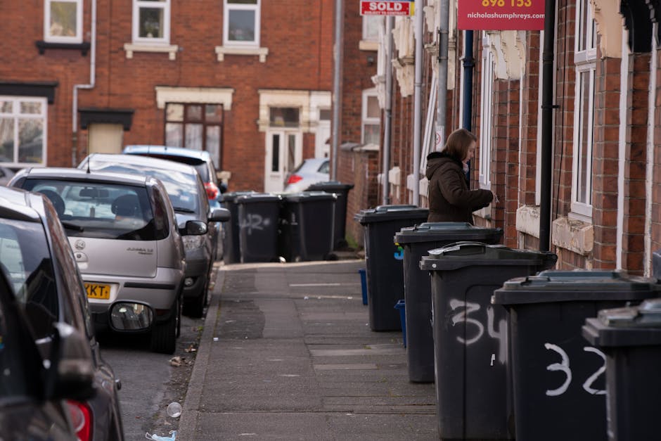 A narrow residential street with parked cars along the left side, including a silver hatchback and a dark-colored vehicle, on a pavement with visible wear and small scattered debris. On the right side, a row of black wheelie bins with white graffiti markings is lined up against red-brick terraced houses with white window frames and decorative stone sills. A woman dressed in a dark hooded coat is standing in front of one of the bins, opening or closing the lid. Above her, a red 'SOLD' sign with a website address partially visible is attached to the building. The scene suggests an area where domestic waste collection is carried out privately, possibly as an alternative to council services, fitting within the context of on-site rubbish removal or private waste disposal. The natural lighting indicates daytime, with a subdued atmosphere typical of urban residential environments where waste is prepared for collection or disposal.