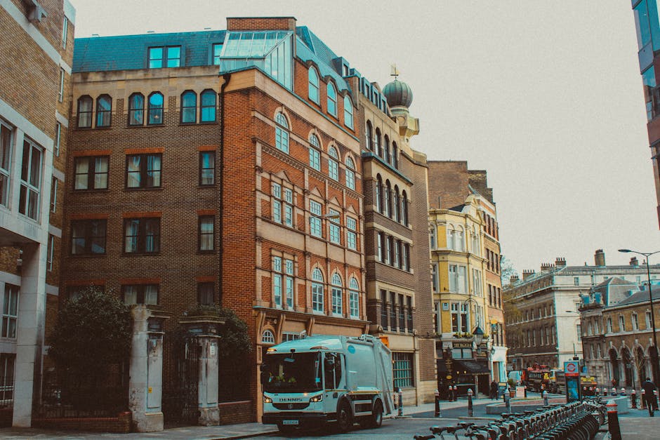 A street scene featuring a variety of multi-storey historic and modern buildings with brick facades, large arched and rectangular windows, and decorative architectural details. In the foreground, a grey waste collection vehicle is parked on the pavement, positioned near the curb, with its side and front visible. The vehicle has a boxy shape with a smooth metal surface, clearly designed for waste collection purposes, and is surrounded by a few small metal barriers and a row of bicycle racks along the sidewalk. The environment is urban, with a mix of stone and brick building materials and a cloudy sky overhead, indicating an overcast day. The scene subtly relates to private waste management services, possibly indicating on-site rubbish collection tasks, with the vehicle appearing ready for urban rubbish removal or bulk waste clearance in the area. The image offers a professional, neutral view of waste collection in a city setting, suitable for illustrating rubbish removal services by companies like wastecollectionfinsburypark.co.uk.