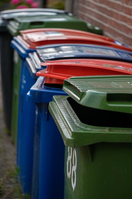 A row of multiple wheelie bins positioned on a pavement against a brick wall, with some bins painted in blue and others in green. The green bins have ribbed, textured lids, while the blue bins have smooth lids. The bins are arranged in a line, with the green bin nearest to the camera in the foreground, and the red, blue, and green lids visible behind. The background features a brick wall to the right and a blurred street scene with grassy areas and other indistinct objects. The scene appears to be outdoors during daylight, with natural lighting illuminating the bins, which are used for waste collection and disposal, possibly as part of an independent rubbish removal service or private waste handling setup managed by a local waste collection company.