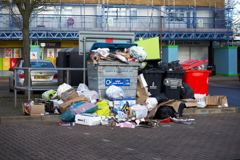 A large pile of mixed household waste and rubbish bags is overflowing from grey and black recycling bins placed on a paved sidewalk area. The waste includes flattened cardboard boxes, plastic bags, paper, and various discarded items, some spilling onto the surrounding pavement. The recyclable materials are visibly crumpled or flattened, with paper documents and cardboard showing clear textures, while plastic bags appear glossy and crumpled. The bins are situated in front of a retail building with storefronts and signage, and a parked car is visible on the left side of the image. An upper floor with exposed metal scaffolding and blue safety netting suggests ongoing construction or maintenance nearby. The scene is captured during daylight hours, with natural light illuminating the area, highlighting the disorderly state of the refuse. This image illustrates a typical scenario involving private waste collection services managing excess rubbish outside commercial or residential premises, reflecting issues with rubbish accumulation that might relate to collection delays or need for external waste removal solutions, such as those offered by [COMPANY_NAME].