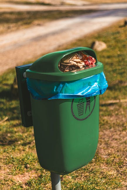 A row of multiple wheelie bins positioned on a pavement against a brick wall, with some bins painted in blue and others in green. The green bins have ribbed, textured lids, while the blue bins have smooth lids. The bins are arranged in a line, with the green bin nearest to the camera in the foreground, and the red, blue, and green lids visible behind. The background features a brick wall to the right and a blurred street scene with grassy areas and other indistinct objects. The scene appears to be outdoors during daylight, with natural lighting illuminating the bins, which are used for waste collection and disposal, possibly as part of an independent rubbish removal service or private waste handling setup managed by a local waste collection company.